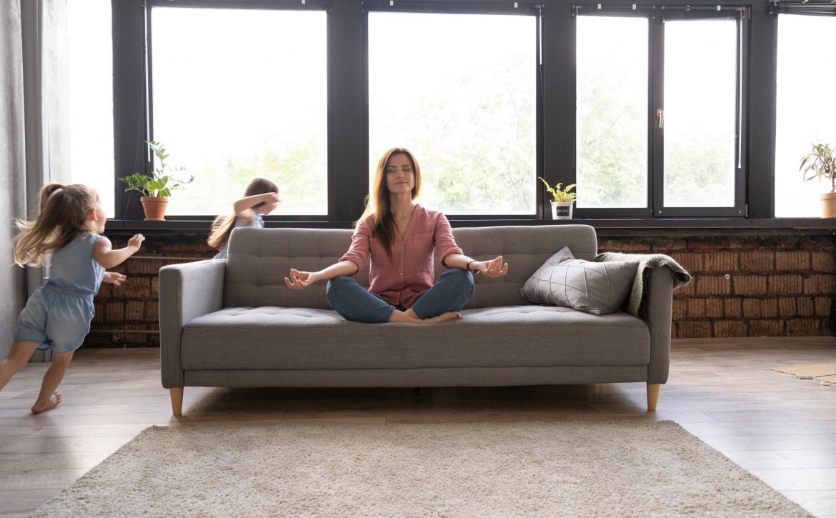 Calm woman concentrating on yoga exercises on couch at home while two noisy kids laughing, running